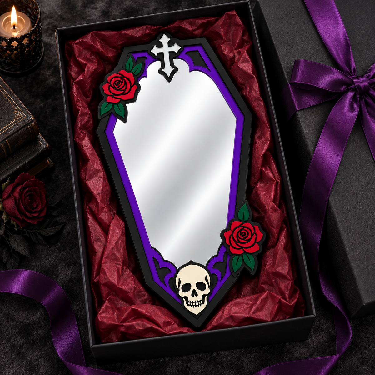 A coffin-shaped mirror with a purple and black frame, decorated with red roses and a skull, presented in a black gift box lined with red tissue paper. A matching black gift box with a purple ribbon is visible on the right, along with a candle and rose on the left.